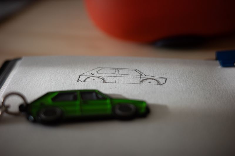 A green VW Golf keychain resting on a notebook next to a pencil sketch of the same car